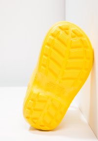 Chaussures jaunes avec un motif de semelle texturé et surélevé. Présentent une forme arrondie et une surface lisse. Logo de la marque visible sur la semelle.