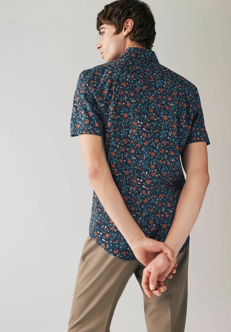 Camicetta a maniche corte, con bottone, di colore blu navy con un motivo floreale in rosso e verde. Design con colletto, retro dritto e vestibilità comoda.