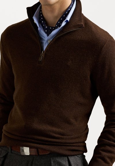 Pulover marrón de punto con cuello con cremallera, llevado sobre una camisa de color azul claro con cuello y lunares blancos, combinado con un cinturón marrón.