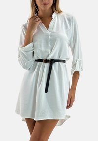 Robe blanche à manches longues avec un col en V, cintrée à la taille par une ceinture noire ornée d'une boucle dorée, fabriquée dans un tissu lisse et léger.