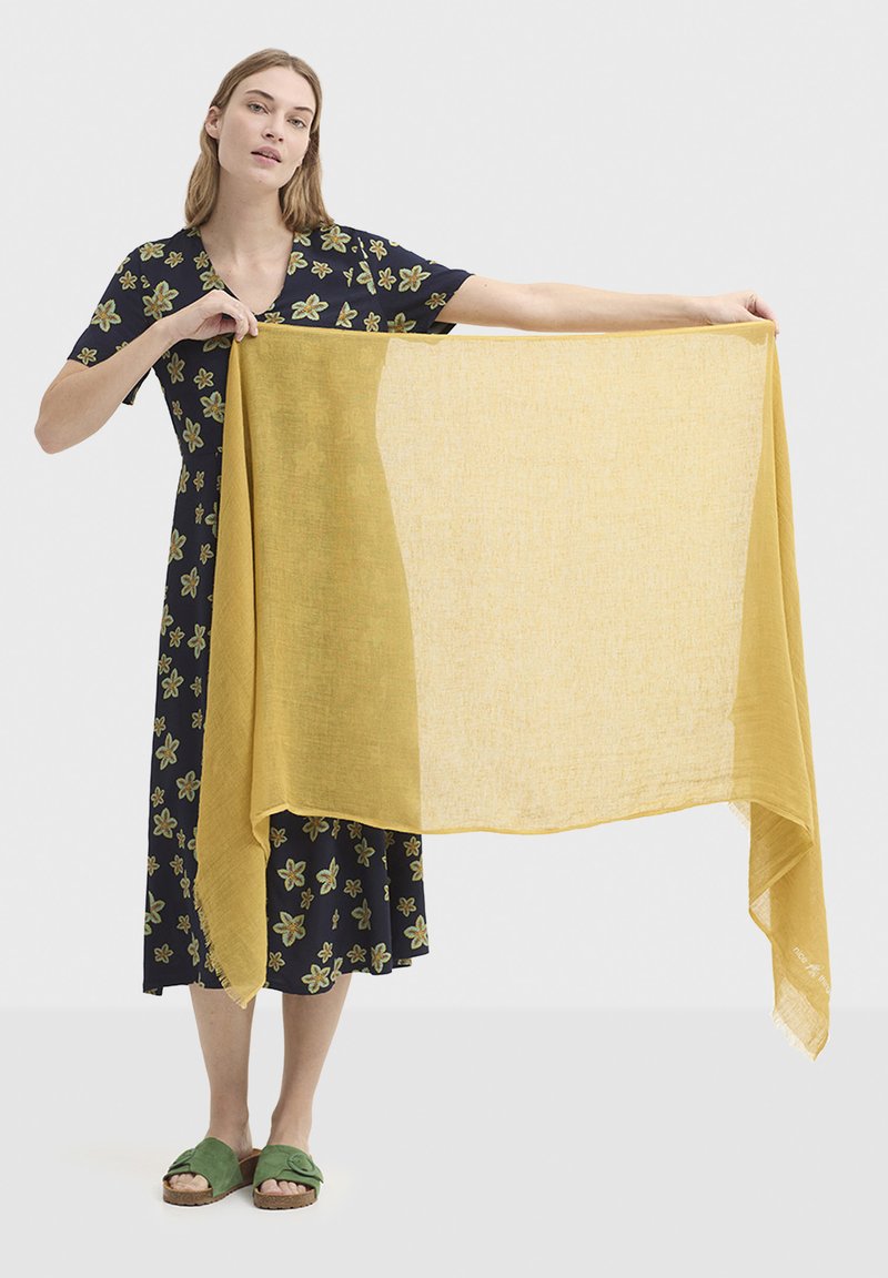 Écharpe légère jaune moutarde tenue par une personne, assortie à une robe fleurie bleu marine et des sandales vertes. L'écharpe a une finition transparente et texturée.