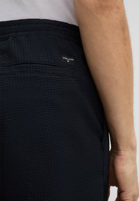 Zwarte textuur shorts met een elastische tailleband, voorzien van een klein logo-label en een achterzak, zijaanzicht weergegeven.