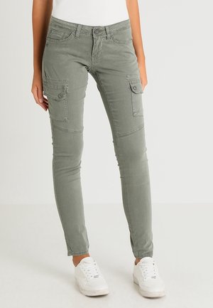 Pantalon cargo vert olive coupe ajustée avec poches à rabats boutonnées sur les cuisses, porté avec des baskets blanches sur un fond clair uni.
