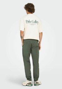 Hombre con cabello rizado de pie de espaldas, vistiendo una camiseta de manga corta color crema con texto verde, pantalones verde oliva y zapatillas beige con detalles verdes.