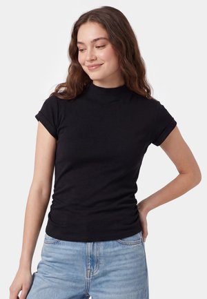 Junge Frau mit langen braunen Haaren, die ein eng anliegendes schwarzes Kurzarmtop und hellblaue Jeans trägt, lächelnd und nach unten schauend.