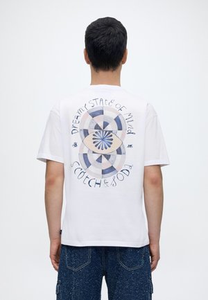 Moški, ki nosi belo majico z modrim in bež krožnim očesnim dizajnom in besedilom "Dreamy state of mind Scotch & Soda" na hrbtu, kombinirano z modrimi hlačami.