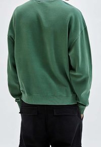 Groene katoen sweatshirt met een ontspannen pasvorm, ribgebreide manchetten en zoom, met afhangende schouders en een zachte textuur. Zwarte broek gedeeltelijk zichtbaar.