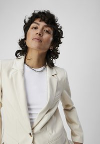Blazer de couleur crème avec une coupe ajustée, fermeture à un bouton, et deux poches avant, porté sur une chemise blanche à côtes, avec des bijoux en argent.