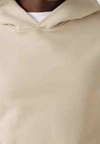 Felpa con cappuccio beige con una finitura morbida e strutturata, dotata di cappuccio con coulisse e colletto di camicia bianca a strati sotto.