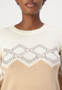 Maglione lavorato a maglia con una sezione superiore color crema e una sezione inferiore beige, caratterizzato da un motivo a rombi arricchito da dettagli in filo metallico.