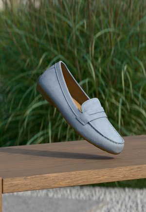 Mocassino in camoscio azzurro chiaro con punta arrotondata, dettagli cuciti e una fascia decorativa sulla parte anteriore, posato su una superficie in legno.