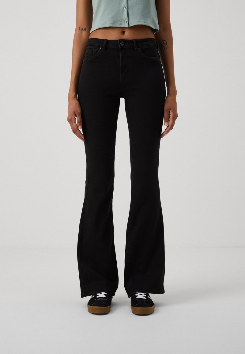 Jean noir évasé à taille haute, fabriqué en denim, avec des poches avant et une fermeture boutonnée classique. Porté avec des baskets noires.