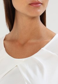 Biała bluzka z okrągłym dekoltem, z fałdami na ramionach i gładkim materiałem, prezentująca czysty i prosty design.