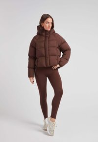 Brązowa krótka kurtka puchowa z kapturem, z pikowanym wzorem i zamkiem błyskawicznym z przodu. W zestawie z dopasowanymi brązowymi legginsami i jasnymi sneakersami.