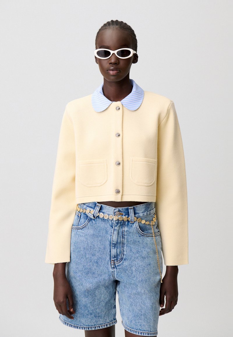 Giacca corta gialla con trama, colletto di camicia azzurro chiaro con bottoni, due tasche frontali e bottoni grigi. Indossata sopra shorts di jeans a vita alta.