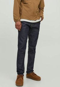Vaqueros de mezclilla azul oscuro con corte ajustado, combinados con una chaqueta ligera marrón, camisa blanca y zapatos de ante marrones.