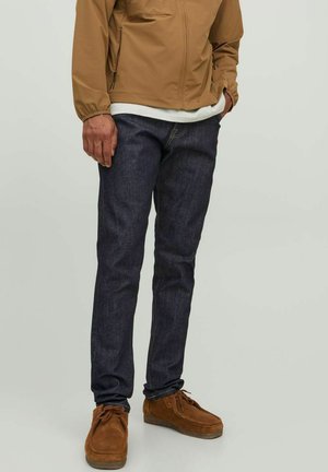 Dark blue denim jeans with a slim fit, paired with a brown lightweight jacket, white shirt, and brown suede shoes.