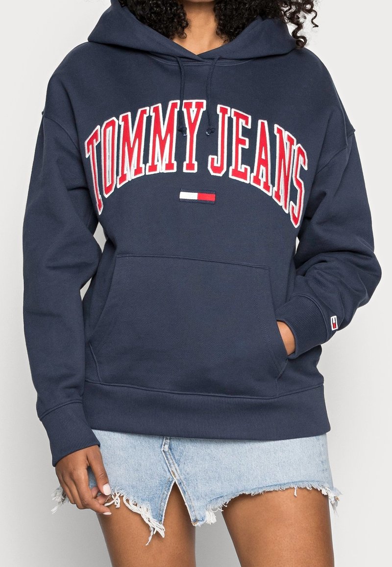 Námořnická modrá mikina s kapucí a červeným a bílým nápisem "TOMMY JEANS". Má přední kapsu, stahovací kapuci a žebrované manžety. Klasický neformální design.