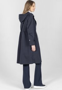 Kobieta ubrana w długi granatowy płaszcz z kapturem, granatowe spodnie i białe trampki, stojąca bokiem na jasno szarym tle.