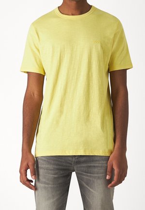 Mann trägt ein schlichtes gelbes Kurzarm-T-Shirt mit kleinem "BOSS"-Logo auf der Brust und verwaschene graue Jeans, steht vor weißem Hintergrund.