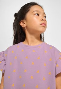 Liliowa bawełniana koszulka z krótkimi bufkami i pomarańczowym nadrukiem motyli na całej powierzchni. Posiada okrągły dekolt i miękką fakturę.