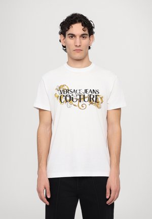 T-shirt bianca in cotone con la scritta "VERSACE JEANS COUTURE" in testo nero e oro, accompagnata da intricati disegni dorati lungo le lettere.