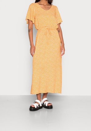 Femme portant une robe midi jaune à imprimé floral blanc, avec des manches courtes volantées, une taille à cordon de serrage et des sandales plateformes blanches.