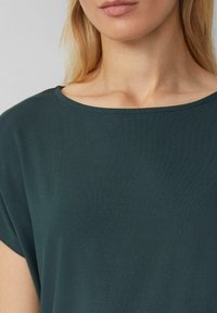 Haut à manches courtes de couleur vert foncé fabriqué dans un tissu doux et lisse. Présente un col large et arrondi et une coupe décontractée.