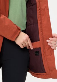 Chaqueta color naranja quemado con forro burdeos, que cuenta con un bolsillo lateral y puños de velcro. La etiqueta interior dice "Reciclado". Textura suave, diseño ajustado.