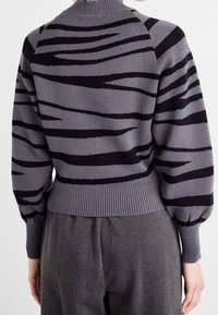 Pull gris avec rayures noires en forme de tigre, col et poignets côtelés, coupe ample, texture douce, et manches légèrement bouffantes.