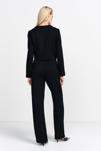 Giacca nera accorciata con colletto di perle testurizzate, abbinata a pantaloni neri aderenti, mostrata da dietro. Tessuto morbido, silhouette snella.