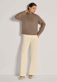 Brauner Strickpullover mit hohem Hals, gerippter Textur und Raglanärmeln, kombiniert mit cremefarbener Cordhose im weiten Schnitt und beigen High Heels.