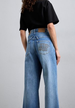 Kvinde iført lyseblå jeans med brede ben, broderet baglomme og en sort kortærmet top, stående foran en ensfarvet grå baggrund.