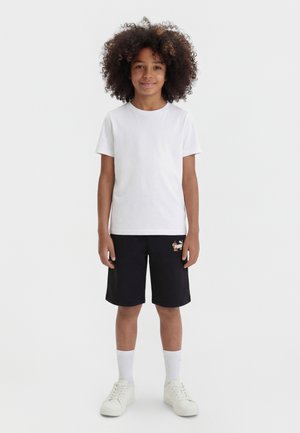Παιδί με σγουρά μαλλιά, φοράει απλό λευκό μπλουζάκι, μαύρο σορτς με πολύχρωμο λογότυπο, λευκές κάλτσες και λευκά αθλητικά παπούτσια, στέκεται σε λευκό φόντο.