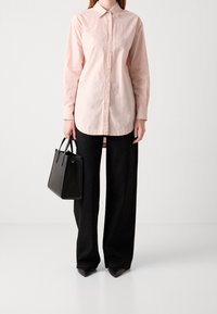 Kvinna klädd i en ljusrosa skjorta med knappar, svarta vida byxor med subtila prickar, svarta spetsiga skor, och håller en svart handväska.