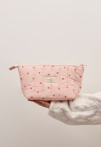 Quiltet, rosa kosmetikkveske med små røde hjerte mønstre, med glidelås på toppen og merke som leser "THE FLAT LAY CO."