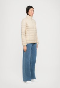 Vrouw staat in een beige gewatteerde jas, wijdvallende blauwe jeans en witte sneakers tegen een plain witte achtergrond, iets naar rechts gericht.