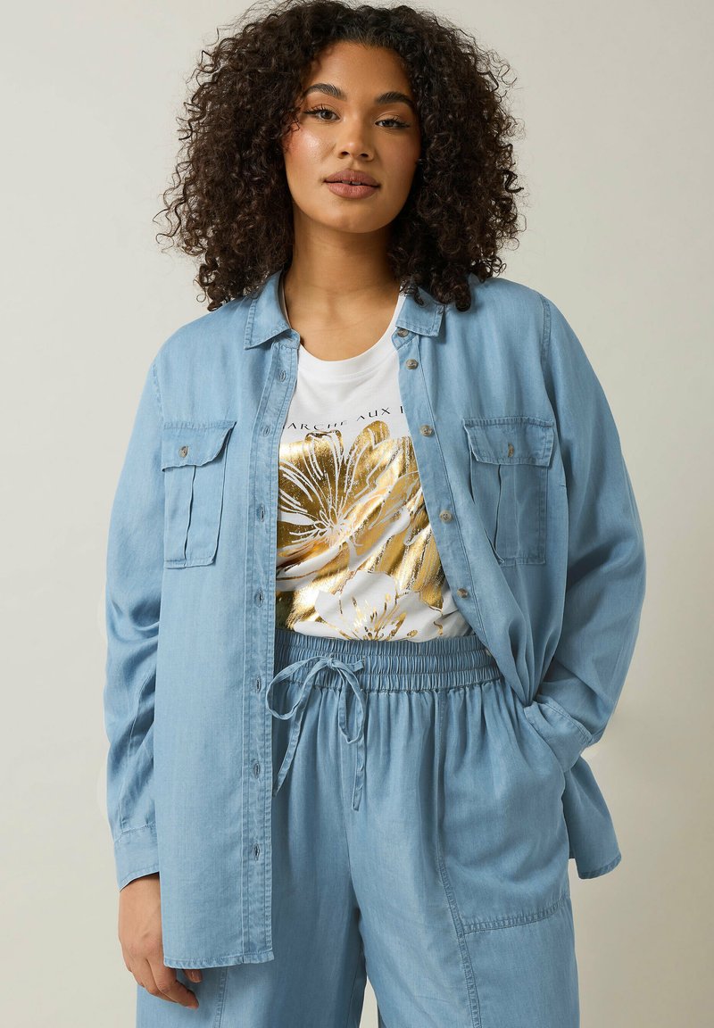 Camicia azzurro chiaro con bottoni, dotata di due tasche sul petto, indossata sopra una maglietta bianca con un motivo floreale dorato, abbinata a pantaloni blu coordinati.