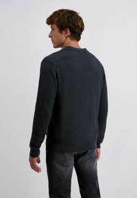 Man in een donker, gestructureerd, lang mouwensweater en vervaagde zwarte jeans, staand met zijn gezicht naar links tegen een effen lichte achtergrond.