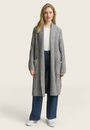 Grijze gebreide cardigan met een lange, open ontwerp, textuurpatroon en twee voorzakken. Gecombineerd met een witte top en blauwe wijde jeans.
