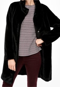 Sort faux fur-frakke med en løs pasform, knaplukning og brede ærmer. Laget over en mønstret bluse med maroon og houndstooth-design.