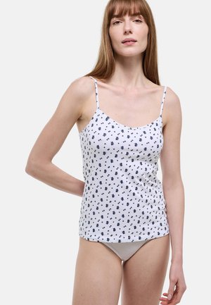 Femme aux cheveux bruns lisses portant un caraco blanc à motif floral noir et une culotte côtelée beige, debout devant un fond uni.