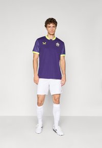 Jeune homme portant un maillot de football violet avec des bordures néon, un short blanc, des chaussettes blanches et des crampons, devant un fond clair.