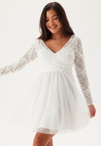 Robe blanche avec de longues manches en dentelle, un décolleté en V et un corsage ajusté, se transformant en une jupe en tulle fluide. Motif floral en dentelle sur le corsage.