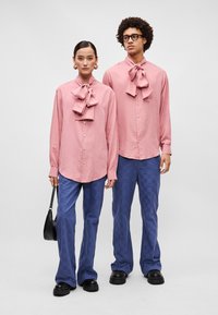 Camisas rosa de botão, com gravatas borboleta oversized, combinadas com calças azuis estampadas. Ambos usam calçado preto e acessórios.