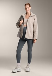 Flisasta jakna v svetlo sivi barvi z visokim ovratnikom, v kombinaciji z temnosivimi leggins in svetlo sivimi športnimi čevlji. Gladka tekstura po celotni površini.