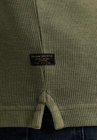 Olivegrüner Strickstoff mit strukturierter Oberfläche, ausgestattet mit einem Etikett, auf dem "FREEDOM UNLIMITED" in goldenen Buchstaben nahe dem Saum steht, sowie einem einzigartigen Belüftungsdesign.