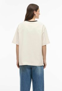 Ženska z dolgimi rjavimi lasmi, oblečena v ohlapno belo majico s črnim ovratnikom in širokimi modrimi kavbojkami, fotografirana od zadaj na belem ozadju.