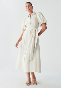 Beige knoopjurk met pofmouwen, stoffen riem in de taille en een lange A-lijn rok. Gladde textuur en lichtgewicht materiaal.