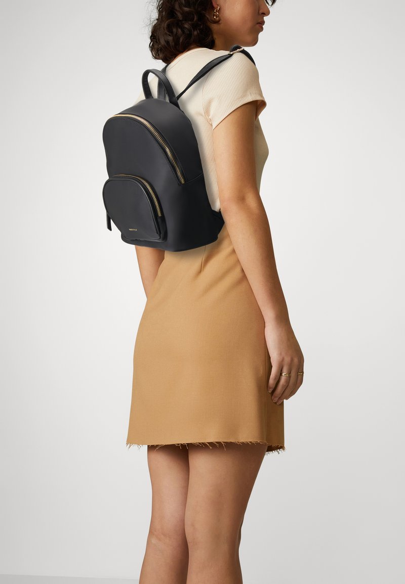 Black leather backpack with curved shape, gold zippers, and subtle branding. Worn on back, paired with a beige dress with frayed hem.